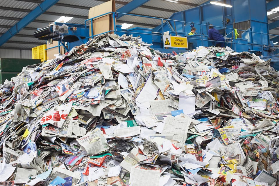 A pile of newspapers in a recycling facilty dwarfs a few workers in the background.
